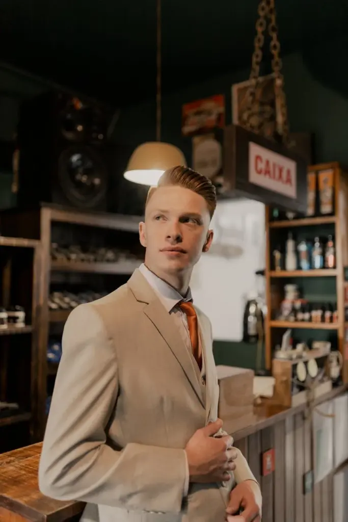 Young man in beige suit with red tie standing confidently in a vintage-style bar.