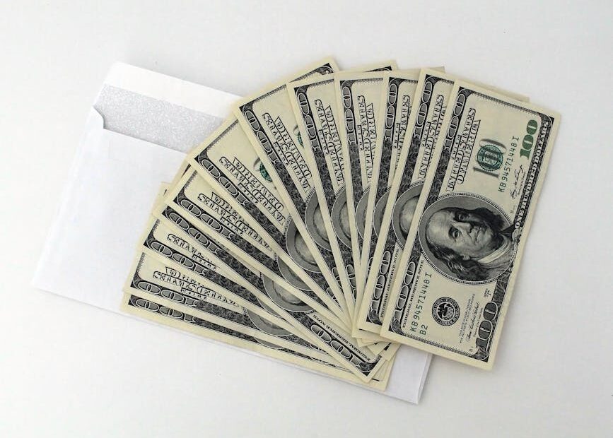 Fan of US $100 bills partially out of a white envelope on a white background.