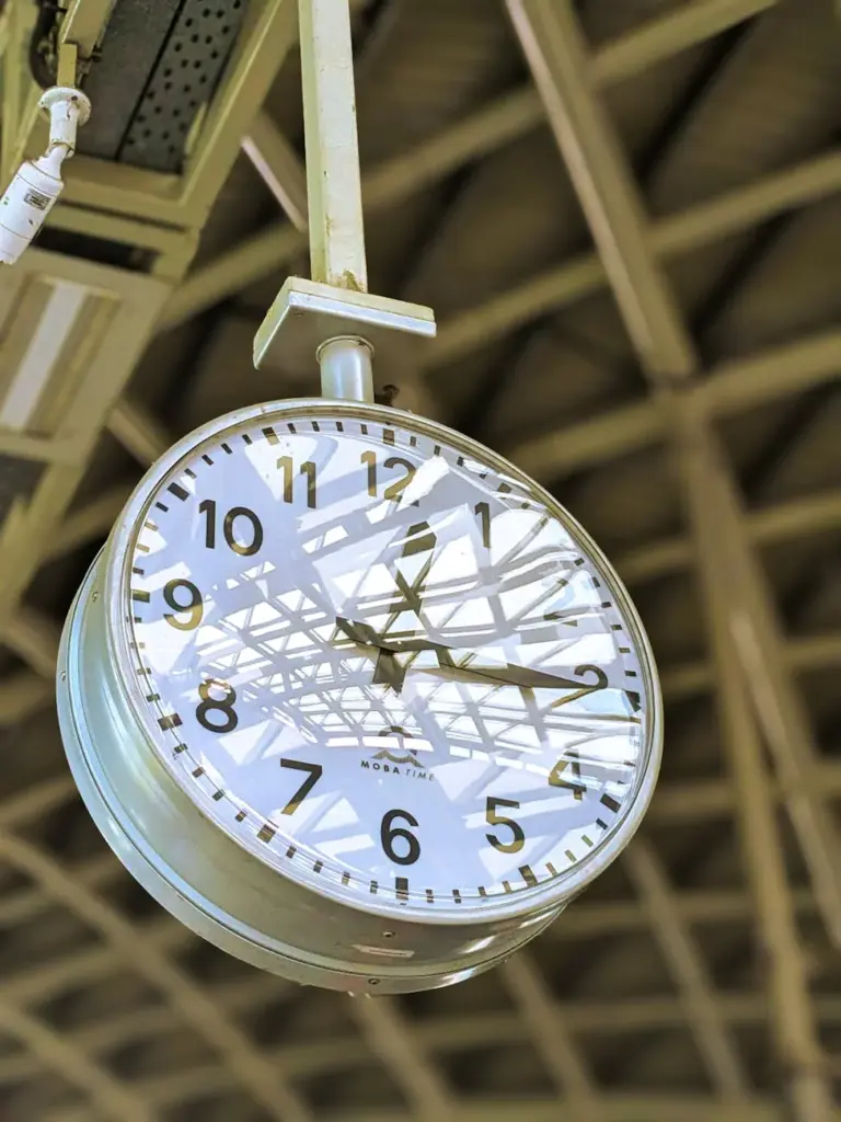 A large vintage clock hanging in a train station, reflecting architectural beams.
