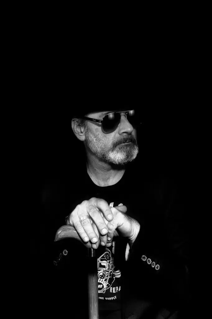 Black-and-white portrait of a senior man in sunglasses and hat against a black background.
