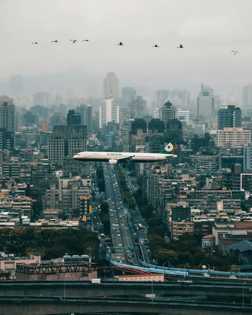 A passenger airplane flying over Taipei's skyline with modern urban buildings and cloudy skies.