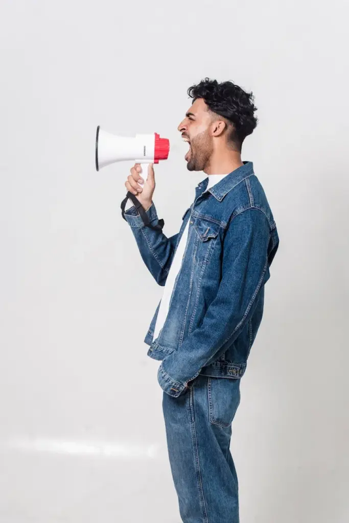 Side view of a man wearing denim, shouting into a megaphone against a white background.