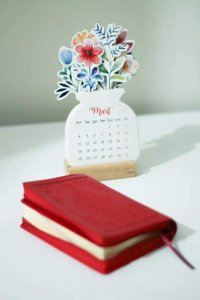 A beautifully designed March calendar with flowers on a white table beside a red notebook.