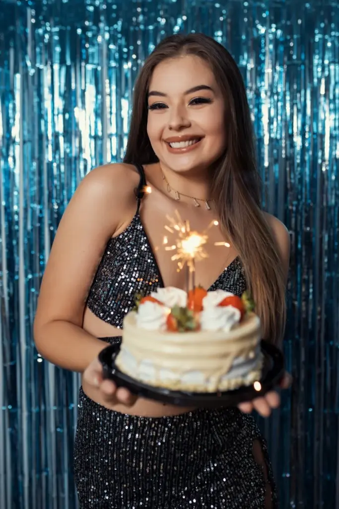 Smiling young woman holding a birthday cake with sparklers in a festive indoor setting.