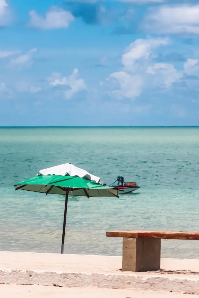 Peaceful beach view in Brazil with green umbrella, bench, and boat on turquoise ocean under blue sky.