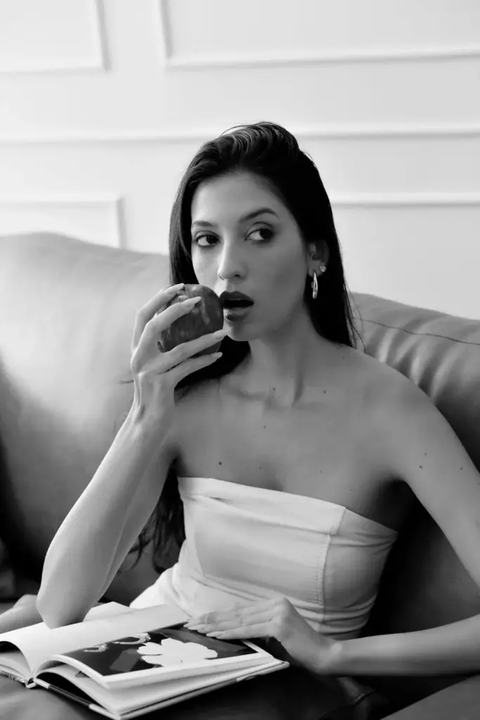 Elegant woman enjoys an apple on a couch, captured in classic black and white.