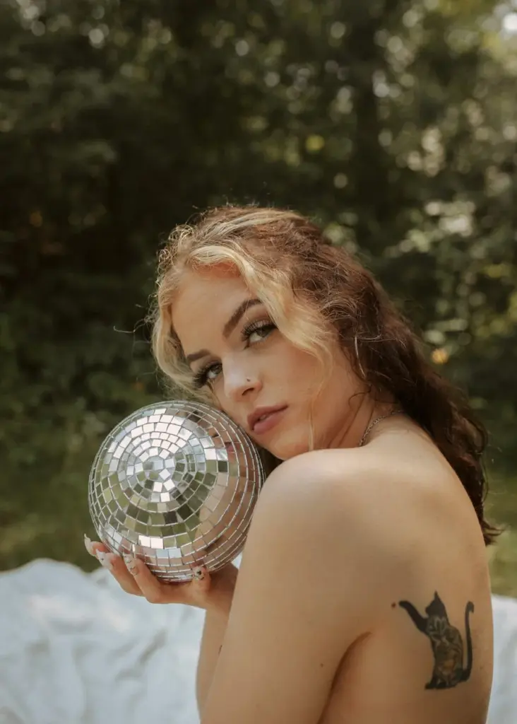 A woman poses artistically with a disco ball outdoors in a lush forest setting.