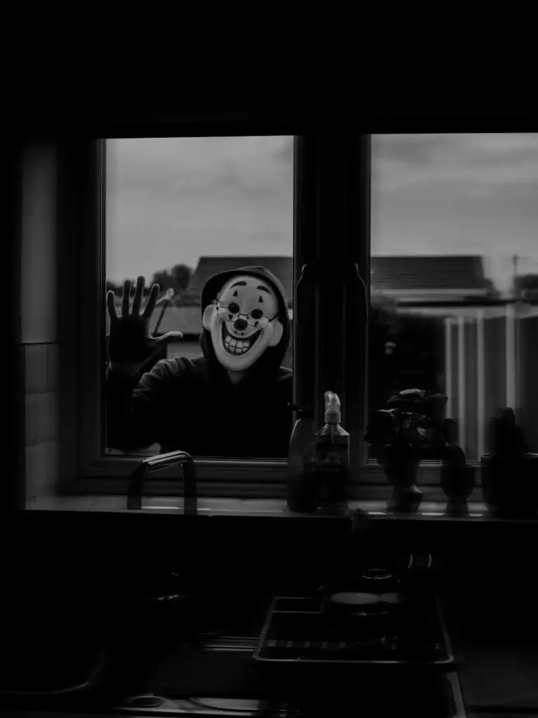 A spooky clown mask peering through a kitchen window in monochrome.
