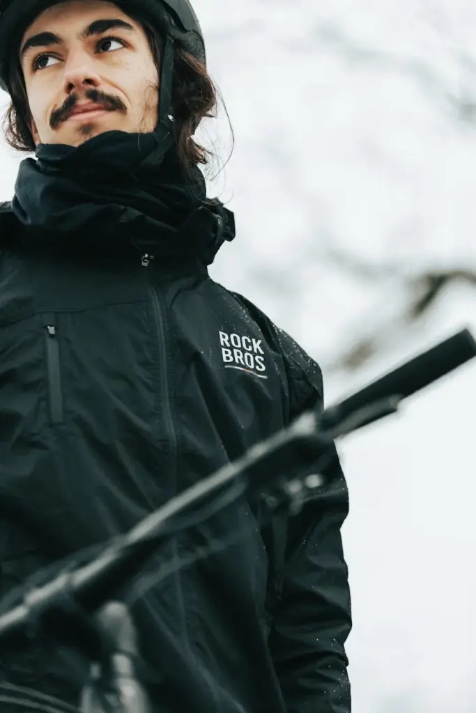 A cyclist in winter gear stands with his bike, looking upward on a snowy day.