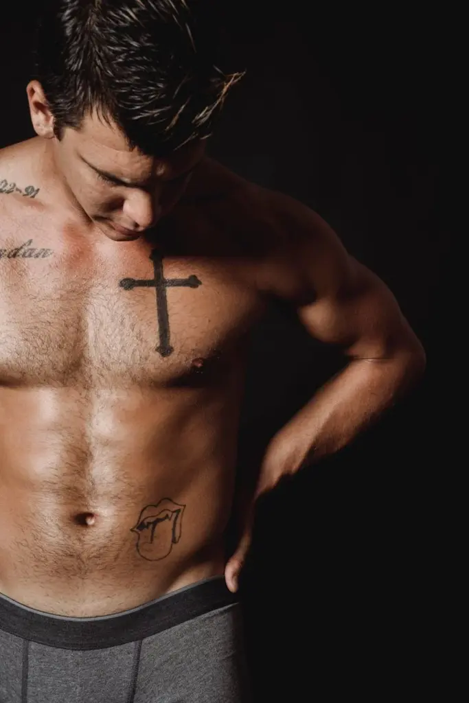 A fit, topless man with distinctive tattoos poses in a studio with a black backdrop.