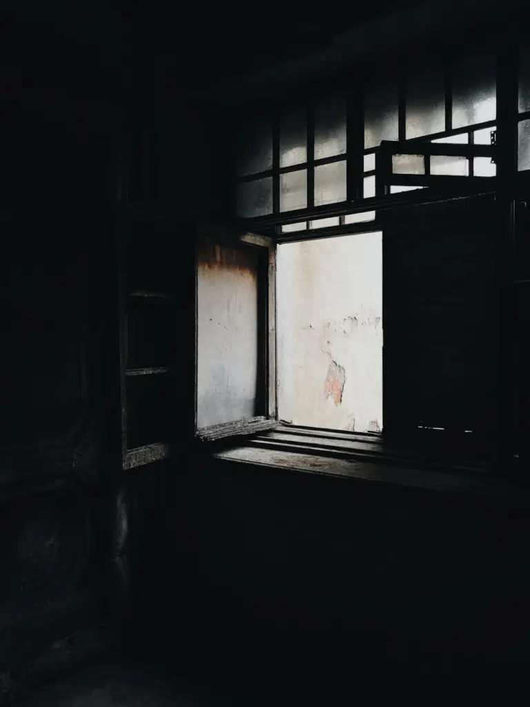 Dimly lit abandoned room with an open window casting eerie shadows.