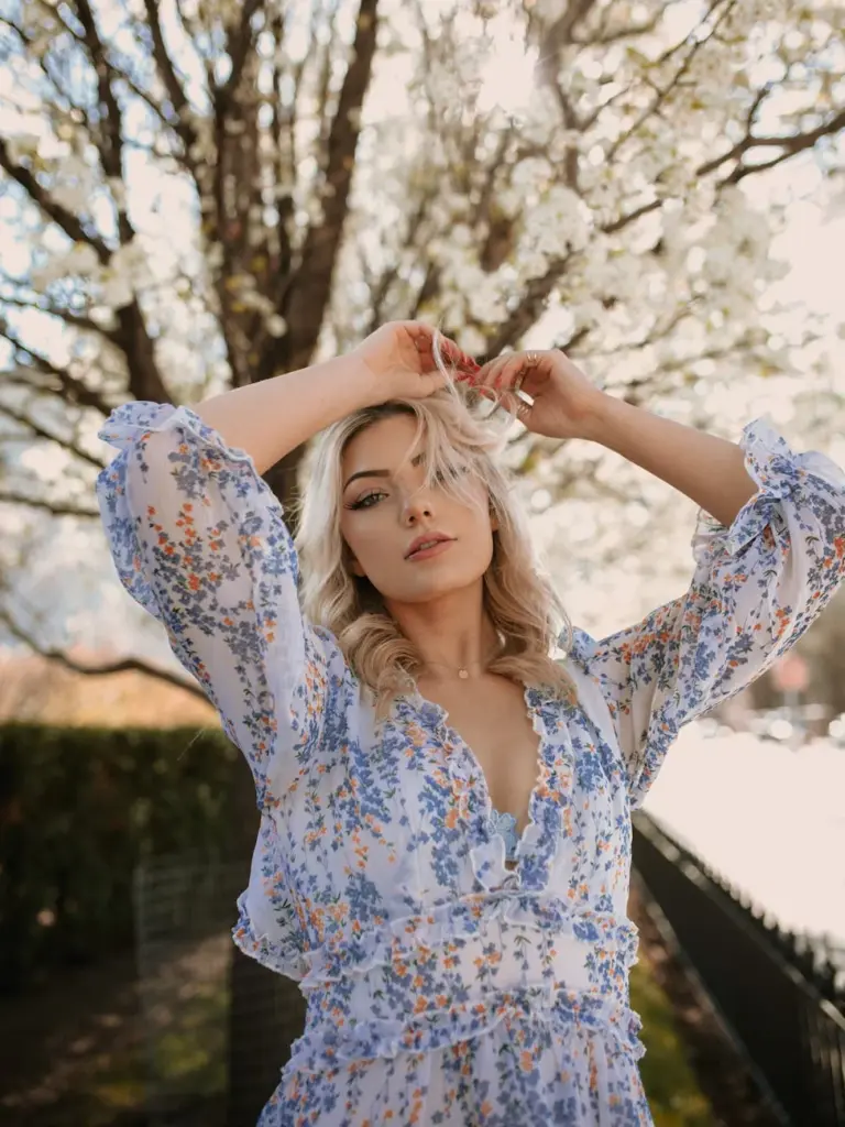 A blonde woman posing in a floral dress surrounded by blooming trees during springtime.