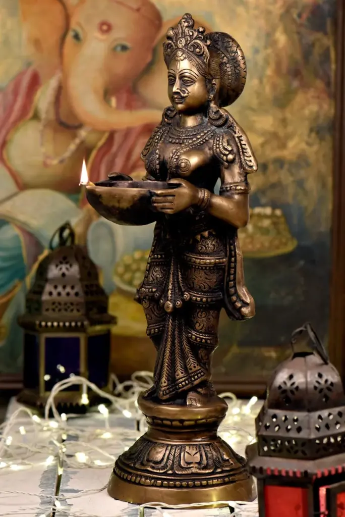Detailed brass Lakshmi statue holding a lit diya, symbolizing prosperity, with Ganesha painting in background.