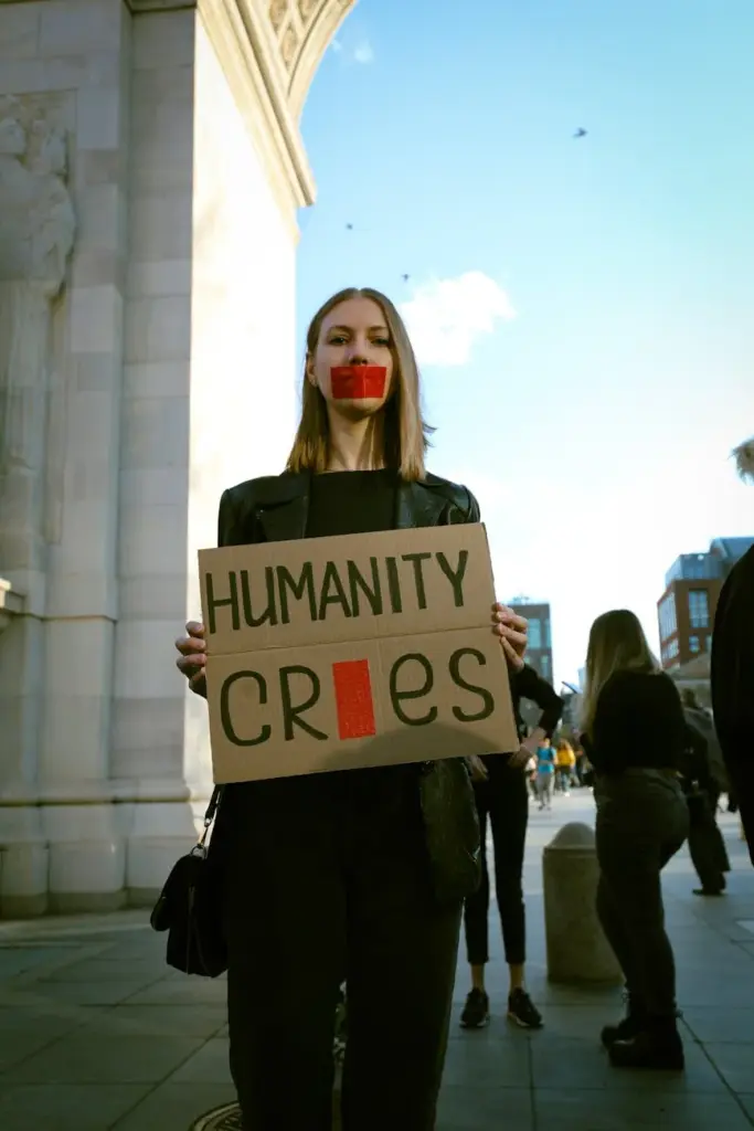 Woman with taped mouth holds protest sign reading 'Humanity Cries' during urban demonstration.