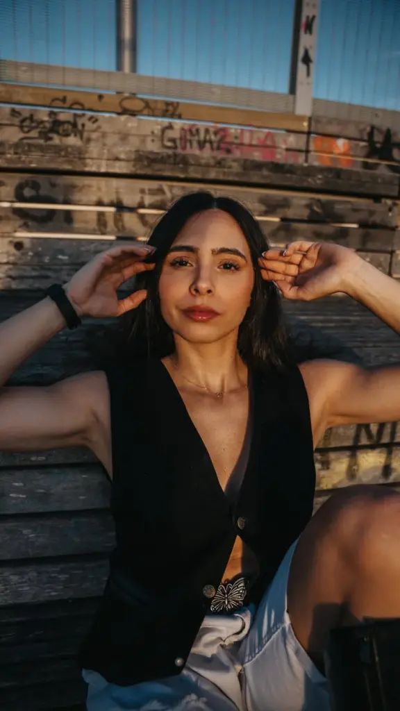 Stylish woman in a black vest sitting outdoors, adjusting hair and enjoying the sun.