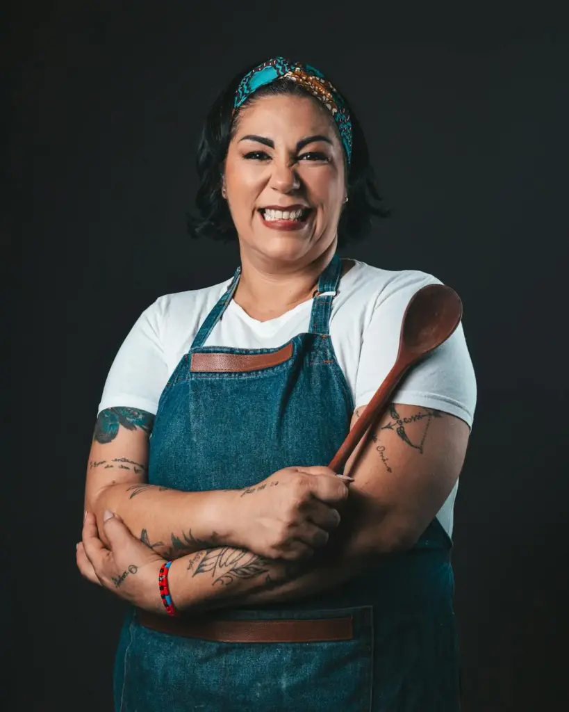 Portrait of a cheerful tattooed chef in apron holding a wooden spoon against a gray background.