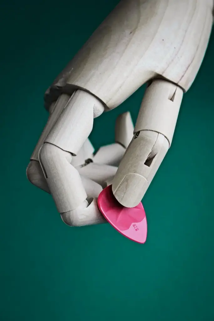 Close-up of a wooden mannequin hand holding a pink guitar pick against a green background.
