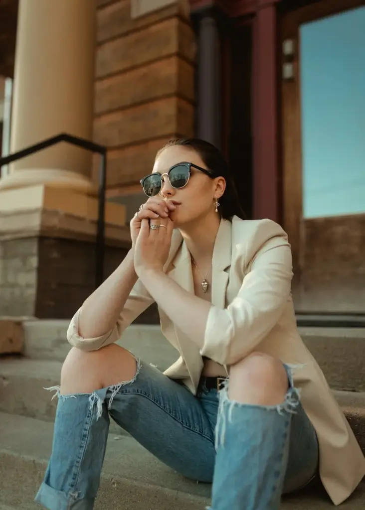 Caucasian woman with sunglasses sitting stylishly on steps wearing a blazer and ripped jeans in Des Moines, IA.