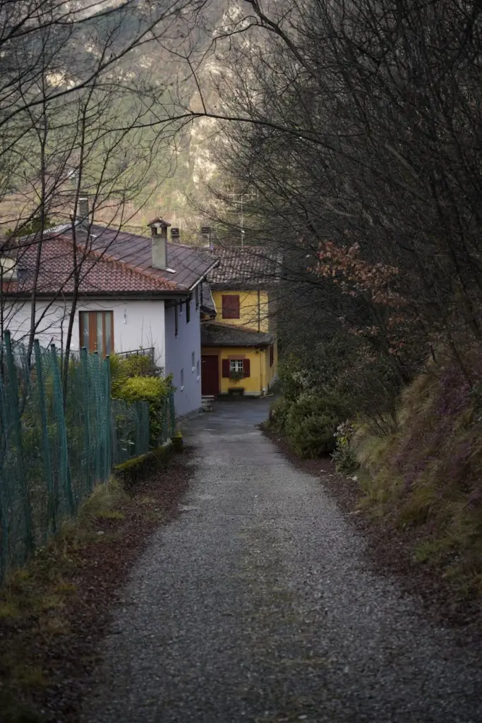 A tranquil path leads through a picturesque village in Lombardia, Italy, with misty mountains in the background.