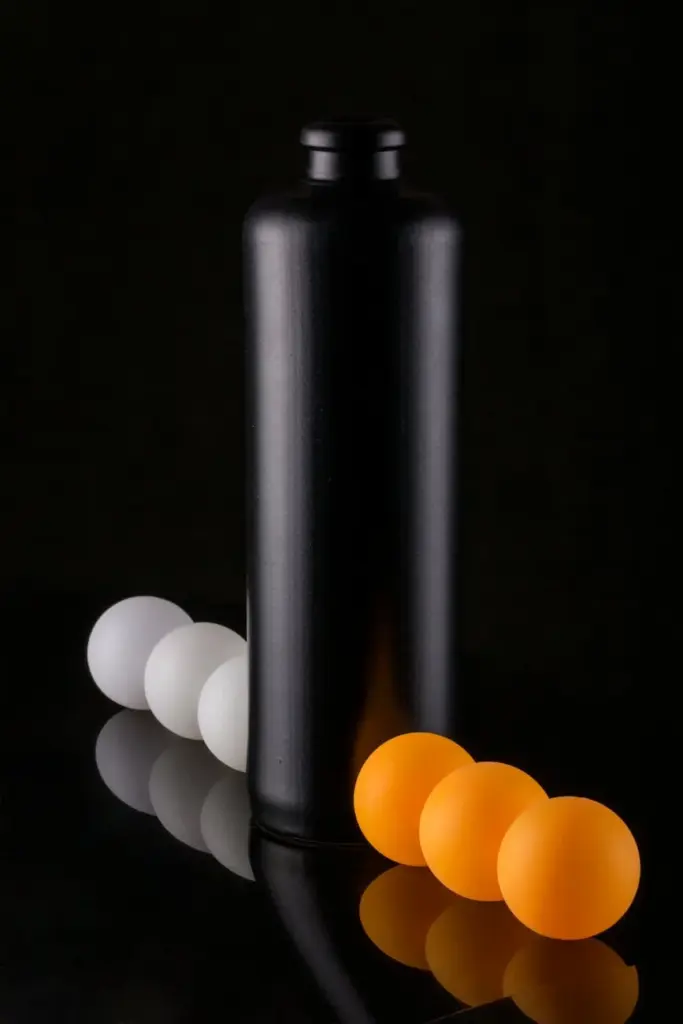 Stylish composition of a black bottle with orange and white ping pong balls on a reflective dark surface.