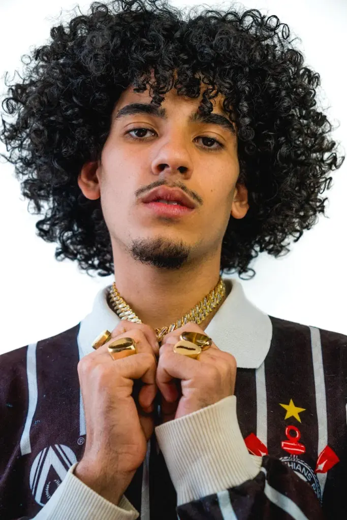 Portrait of a stylish young man with curly hair, wearing gold jewelry and a sporty jacket.