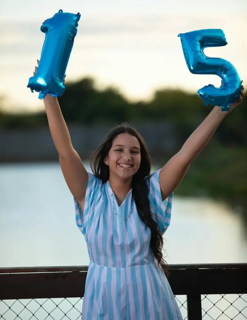 Happy teenager holding blue '15' balloons, celebrating outdoors with a smile.