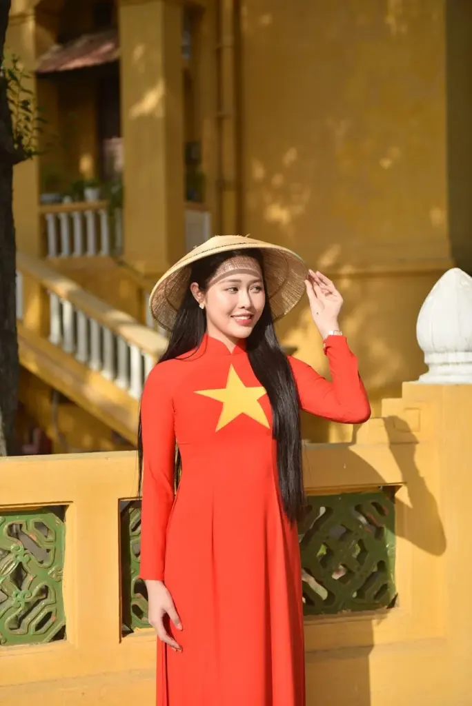 A woman in a red ao dai with a star design stands in sunlight wearing a conical hat.