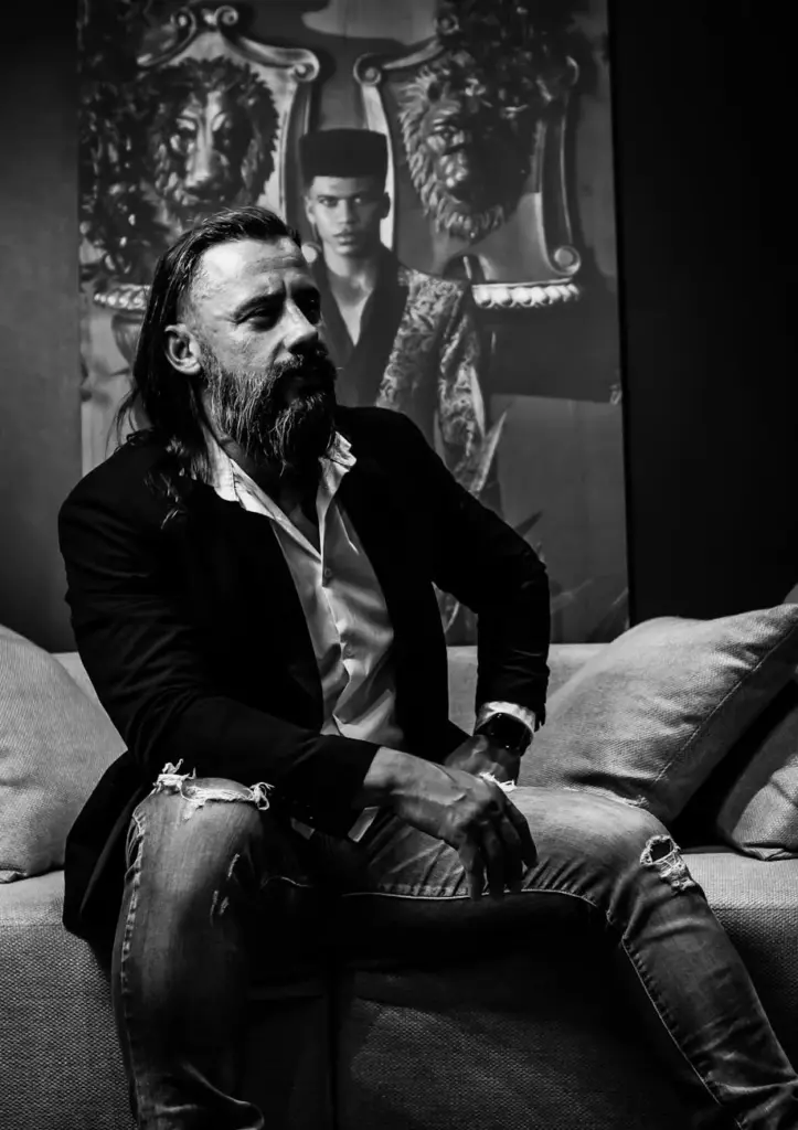 Bearded man in a blazer sits on couch, blending modern style with classic portrait.