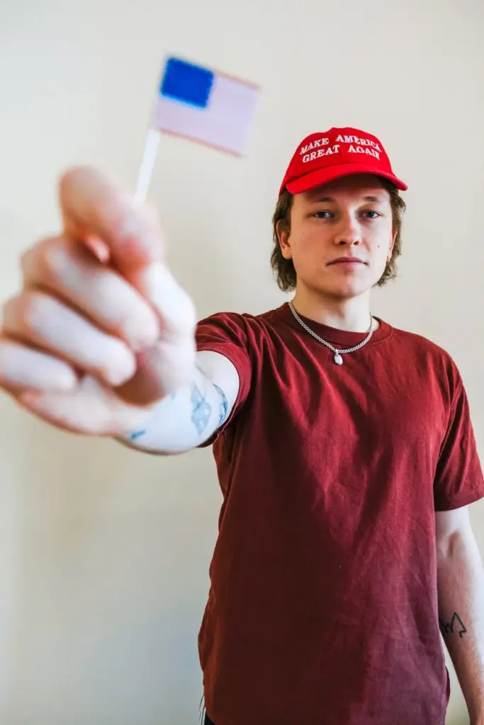 Young adult holding small American flag, wearing a red political hat indoors.