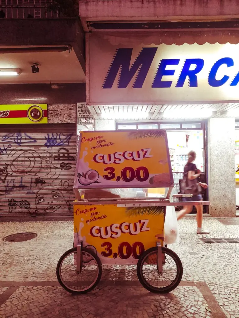 Cuscuz vendor cart in front of a store at night, offering street food in urban setting.