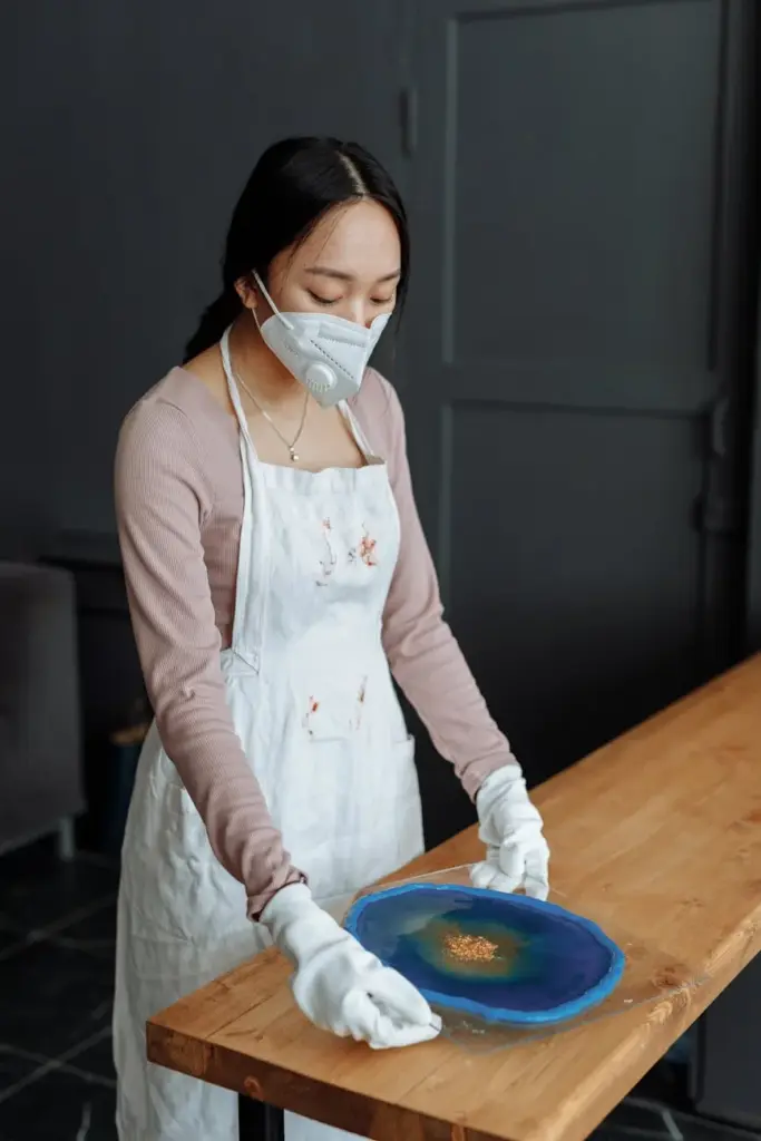 Artist in face mask and apron crafting resin art on wooden table.