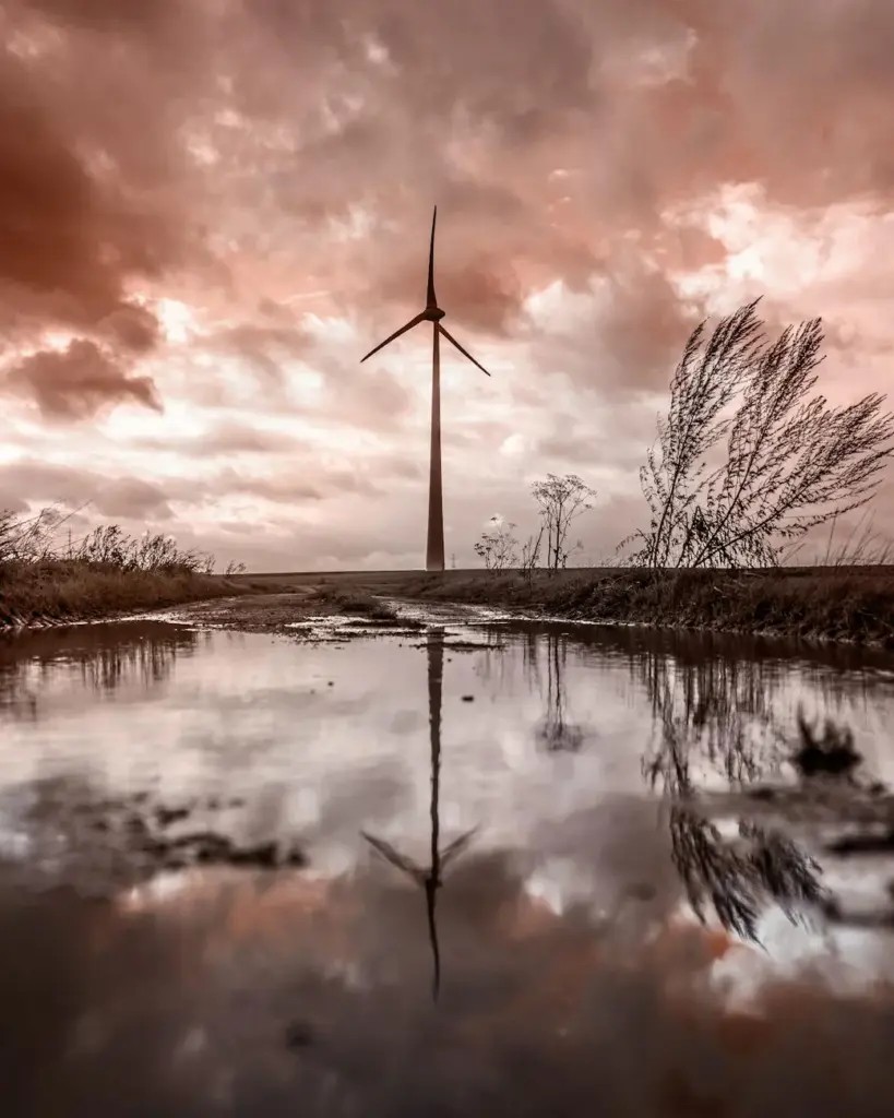 Silhouette of a wind turbine reflecting in a puddle under a dramatic sunset sky.