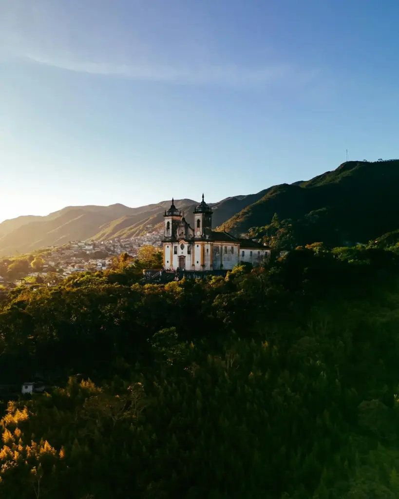 A breathtaking aerial view of a historic church in Ouro Preto, Brazil, surrounded by lush hills at sunset.