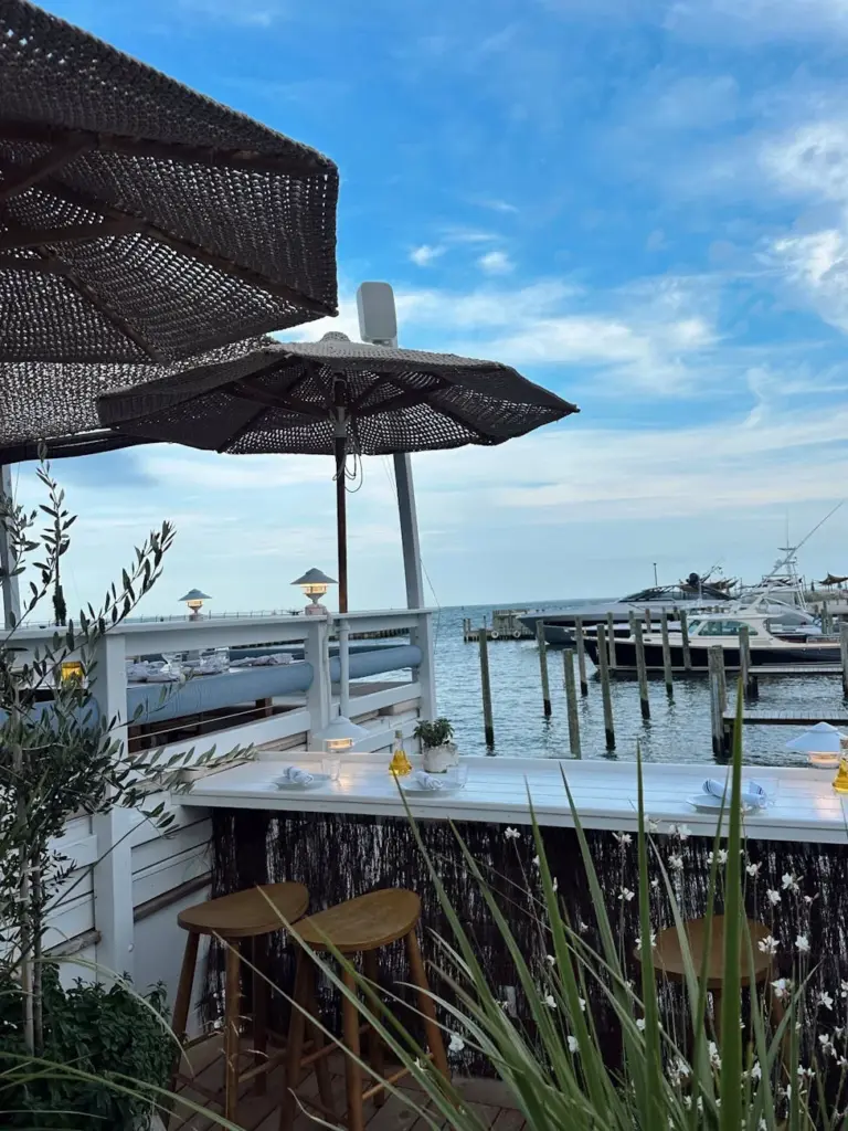 A scenic outdoor bar overlooking a tranquil harbor in Orient, New York with boats and blue skies.