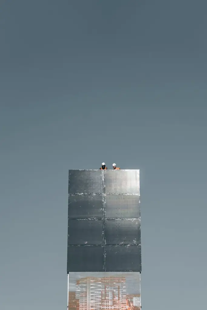 From below of anonymous distant engineers in helmets working on rooftop of modern skyscraper with glass mirrored walls against cloudless blue sky