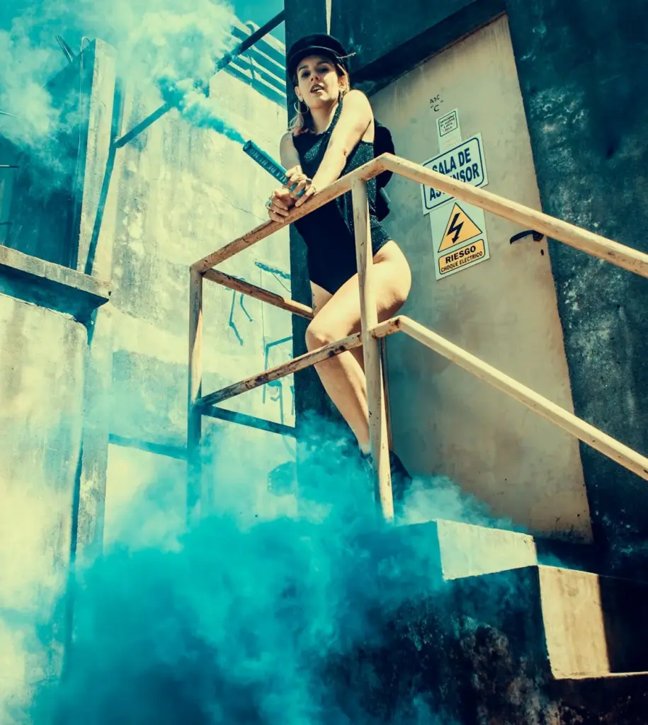 Fashionable woman with smoke bomb on industrial stairs in blue haze outdoors.