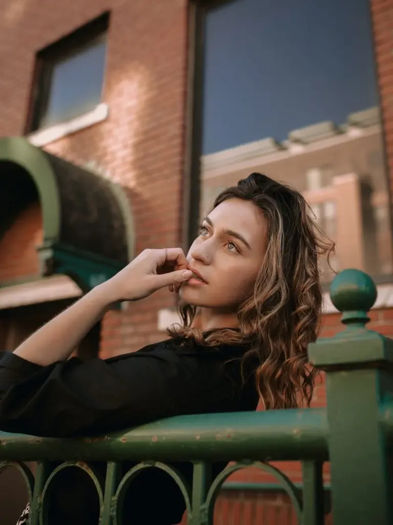 Young woman in black top leaning on a fence with urban building backdrop, showcasing fashion and style.