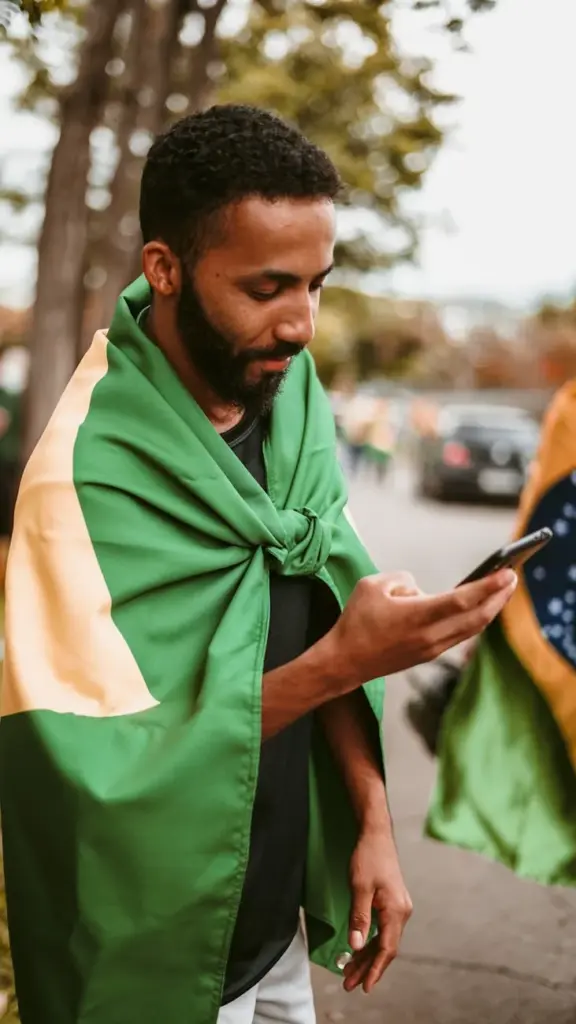 A man outdoors wrapped in the Brazilian flag, looking at his smartphone, showcasing national pride.