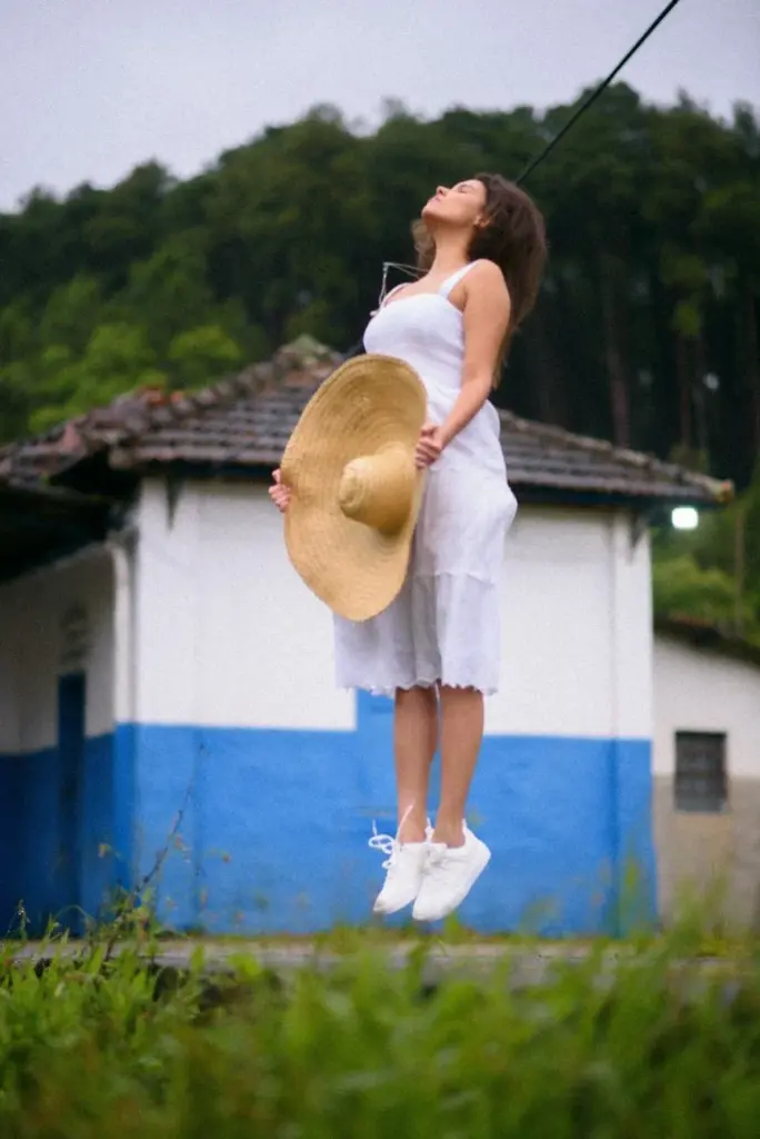 Young woman in white dress jumping outdoors holding a sun hat, set against a rustic building and forest backdrop.