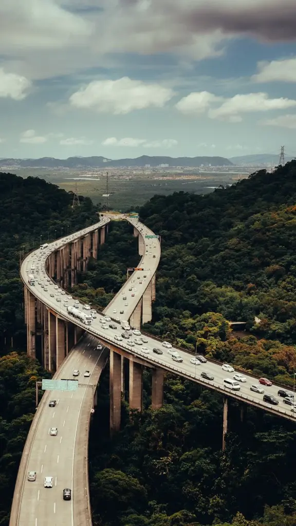 Aerial view of a winding highway through lush forests and hills, with distant cityscape.