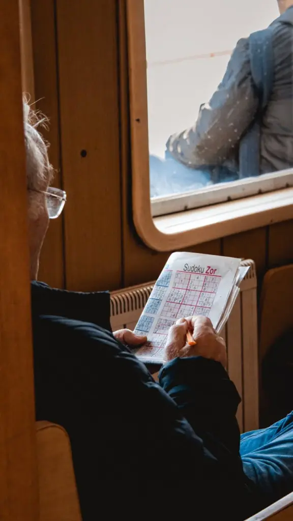 Elderly man enjoys a relaxing moment solving Sudoku puzzle while sitting by a window indoors.