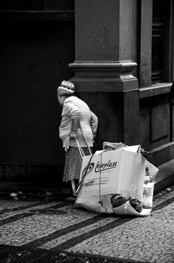 Elderly woman using a walking aid, pulling a cart with cardboard in a city street.