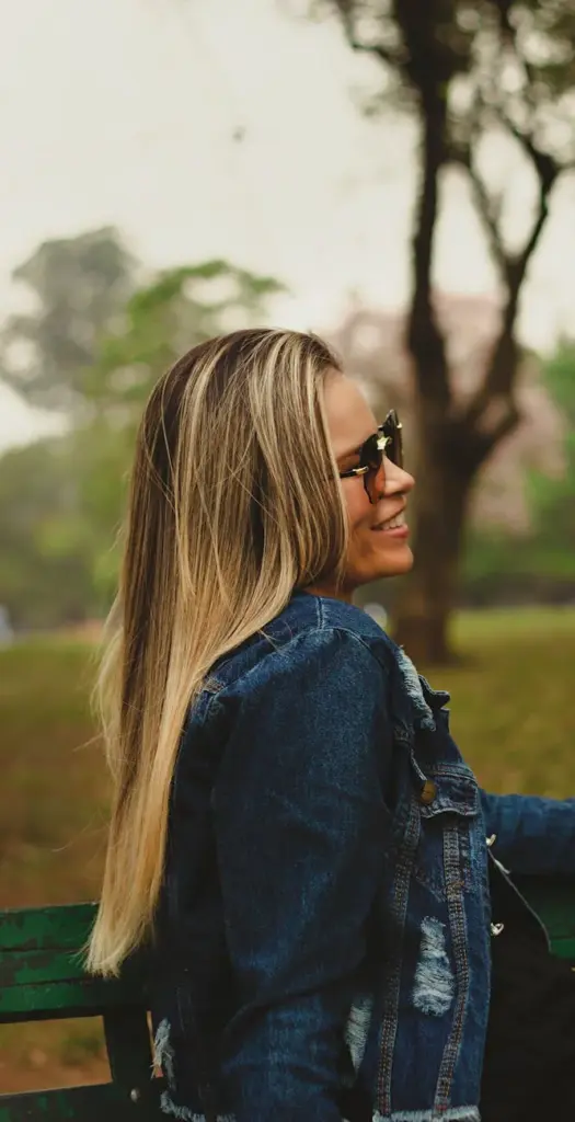 Side view of a smiling woman wearing sunglasses and a denim jacket on a park bench.