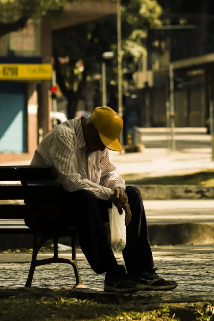 An elderly man with a yellow cap sitting alone on a park bench, looking thoughtful.