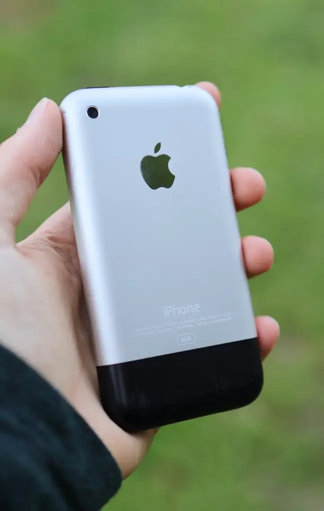 Close-up of a person holding a classic Apple iPhone outdoors on a blurred background.