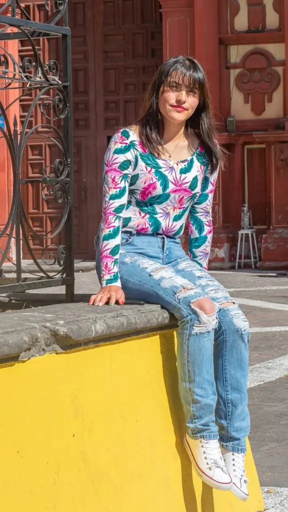 A casually dressed woman sits on a yellow ledge in the sunny streets of Lerma de Villada.