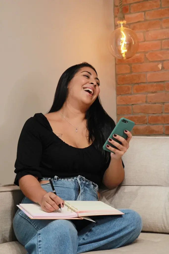 Happy woman laughing while using smartphone and writing in a notebook on a cozy couch.