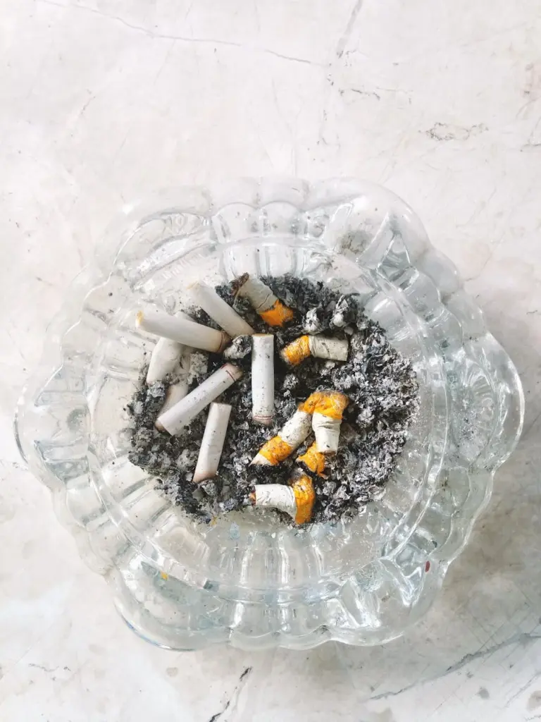 Top view of a glass ashtray filled with cigarette butts and ash on a marbled surface.