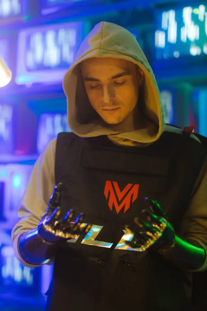 Man in hood with robotic hands in a cyberpunk setting with neon lighting.