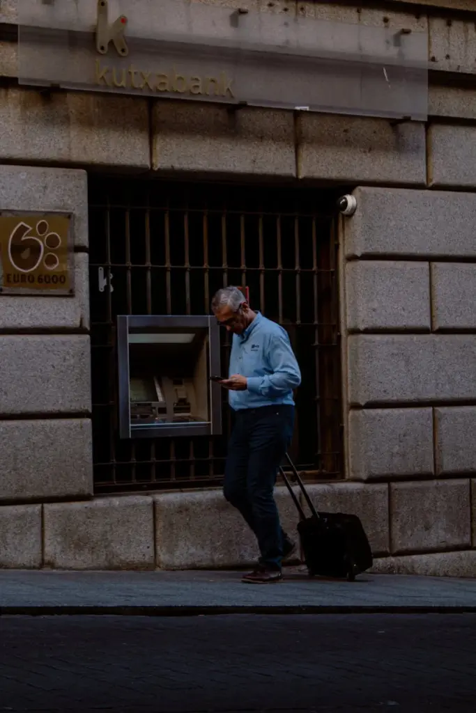 A businessman using an ATM on city streets while checking his phone.